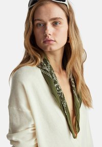 Pletený kardigan v krémové barvě s hlubokým výstřihem do V, vybavený zeleným šálovým límcem se vzorem v bílé barvě. Měkká textura a uvolněný střih.