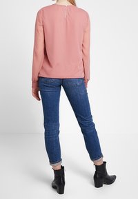 Blusa rosa pallido a maniche lunghe con zip sul retro, abbinata a jeans in denim blu, orlati alla caviglia, e stivali neri alla caviglia.