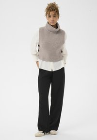 Világosszürke kötött mellény magas, felcsavarodó nyakkal, nagy barna gombokkal az oldalán. Fehér inggel és fekete, bő szárú nadrággal párosítva.