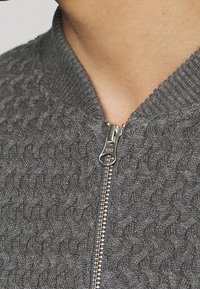 Grå ziptröja med en strukturerad stickad design, med en ribbad krage och en silvrig dragkedja fram.