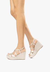 Sandali wedge metallici dorati con cinturini incrociati e punta aperta, dotati di una suola texturizzata in stile intrecciato e fibbia regolabile alla caviglia.