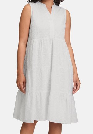 Mouwloze witte jurk tot op de knie met een geborduurd ajourpatroon en een v-hals, gedragen door een persoon met een medium huidskleur tegen een effen achtergrond.