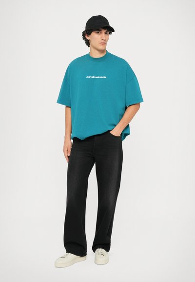 Teal oversized t-shirt med hvid tekst, parret med sorte straight-leg jeans og hvide sneakers. Sort cap med tekstdetalje.