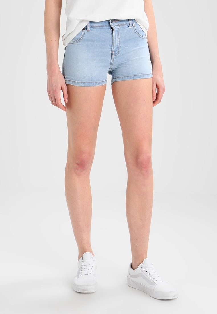 Femme portant un short en jean bleu clair, des baskets blanches décontractées et un haut blanc ample à manches courtes, debout devant un fond uni.
