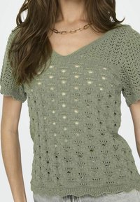 Top de punto de color verde oliva, de manga corta, con escote en V, patrón de tejido abierto y bordes ondulados. Fabricado con un tejido ligero.