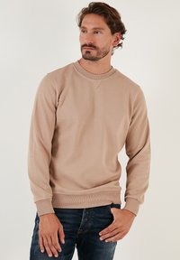 Sweatshirt in cotone beige con maniche lunghe, collo a giro, tessuto testurizzato e polsini e orlo a costine. Indossato con jeans scuri.