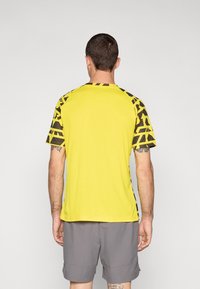 Gul athletic t-shirt med sorte geometriske mønstre på ærmerne, korte ærmer og rund halsudskæring, parret med grå shorts.
