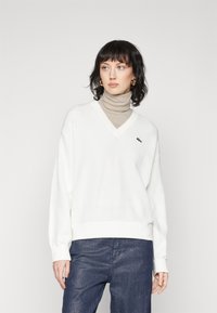 Witte V-hals trui van zacht materiaal met een ribbelrand aan de onderkant en de mouwen; heeft een klein groen logo op de borst, gedragen over een beige coltrui.