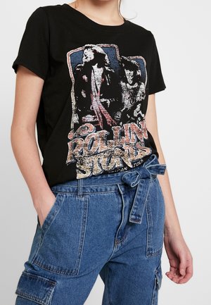Personne portant un t-shirt noir à motif Rolling Stones et un pantalon en jean bleu taille haute avec une ceinture nouée, main dans la poche.