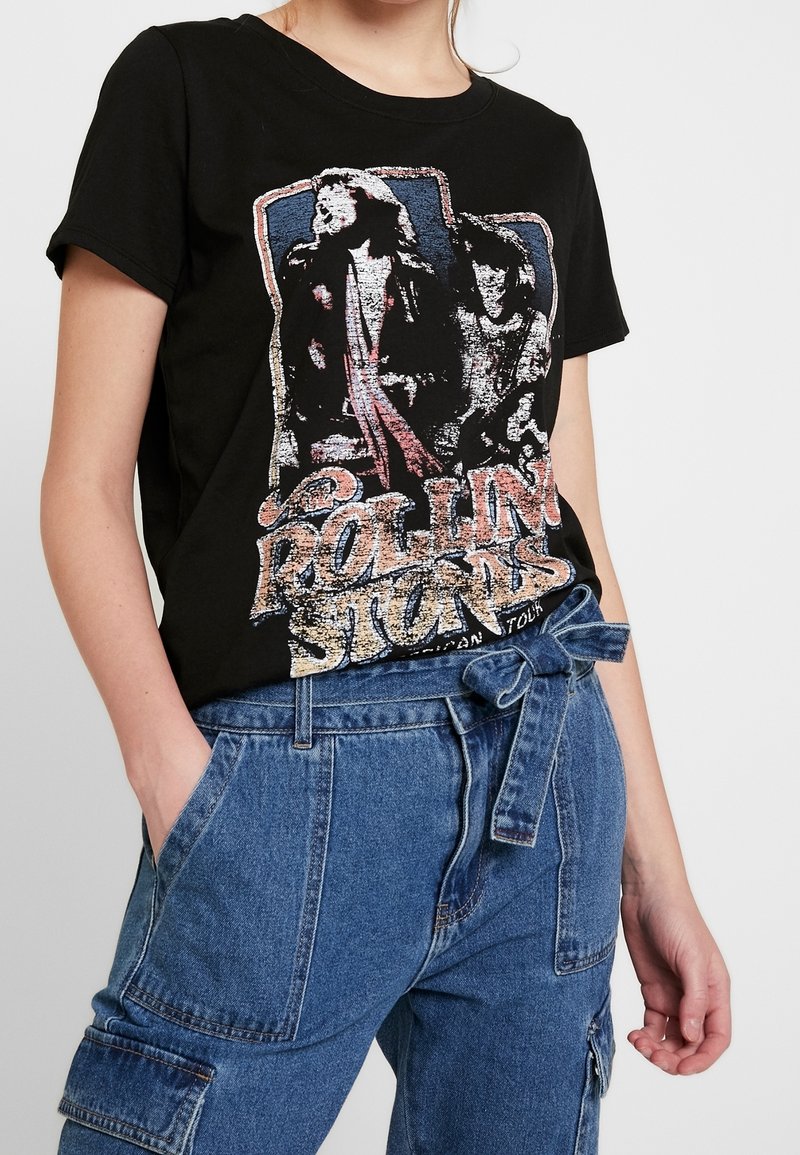 Personne portant un t-shirt noir à motif Rolling Stones et un pantalon en jean bleu taille haute avec une ceinture nouée, main dans la poche.