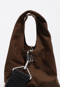 Brązowa torba materiałowa o zaokrąglonym dnie, z dużym uchwytem, chromowanymi elementami i odpinanym czarnym paskiem o strukturalnej fakturze.