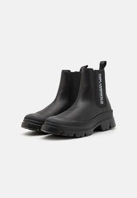 Botas de tobillo de cuero negro con paneles laterales elásticos, suela de goma texturizada y una lengüeta con marca en la parte posterior. Diseño robusto.