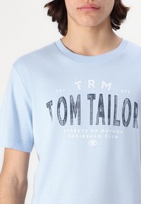 Světle modré bavlněné tričko s krátkým rukávem, s velkým kruhovým grafickým prvkem v tmavě modré barvě a bílým textem. Design obsahuje nápis "TOM TAILOR" a další detaily.