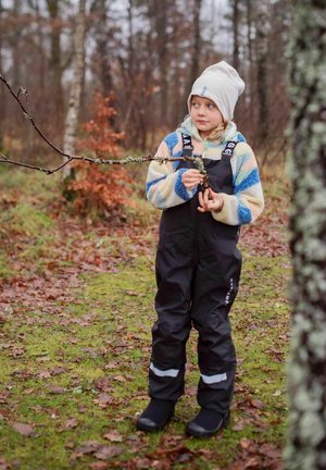 Lapsi, jolla on päällä kuvioitu fleecetakki sinisillä, vaaleanbeigeillä ja oransseilla raidoilla, mustat vedenpitävät haalarit ja mustat saappaat, pitää kädessään oksaa.