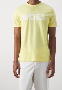 Man wearing a light yellow T-shirt with "BOSS" in white letters, paired with white pants, standing against a plain light background.