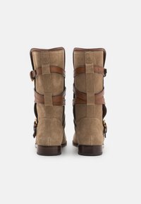 Bottes en daim marron avec des courroies en cuir sur le dessus et un talon en bois. Coutures visibles et détail du logo à l'arrière.