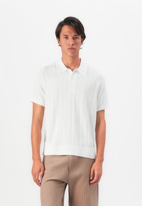 Man wearing white short-sleeve polo shirt with vertical lines and beige pants, standing against a plain white background.