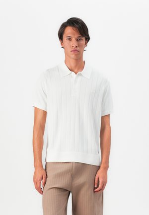 Man som bär en vit kortärmad polotröja med vertikala linjer och beige byxor, stående framför en vit bakgrund.
