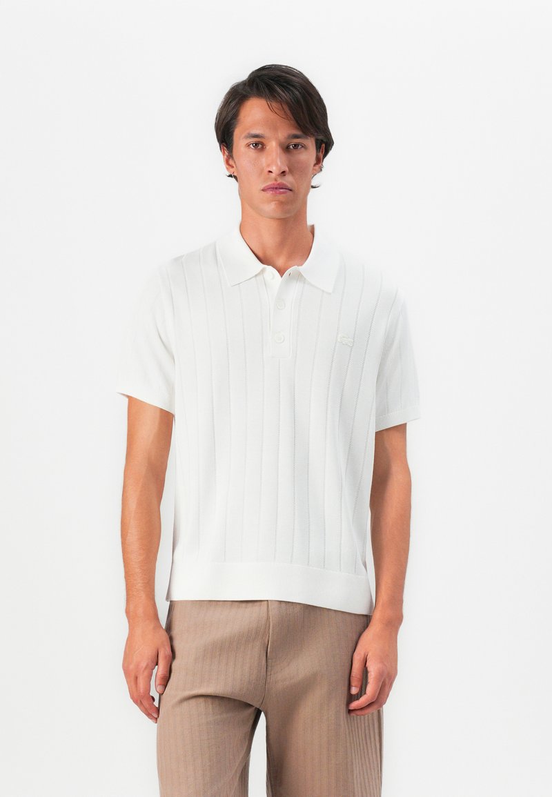 Man wearing white short-sleeve polo shirt with vertical lines and beige pants, standing against a plain white background.