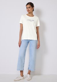 T-shirt crème à manches courtes avec texte bleu, associée à un jean bleu clair à jambes larges et des baskets blanches avec accents beiges ; design simple et épuré.