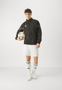 Svart sportjacka med grå detaljer, vita shorts och fotboll. Modellen bär vita strumpor med svarta rand och fotbollsskor.