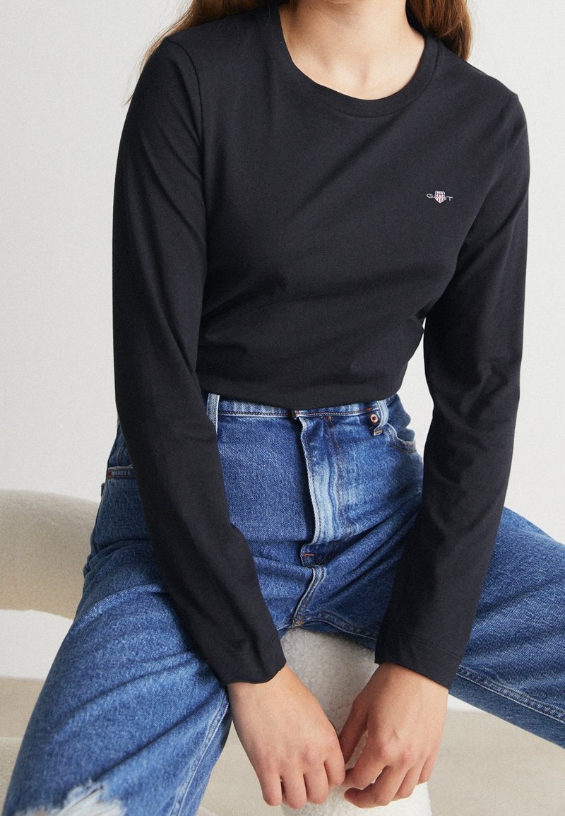 Czarna, long-sleeve t-shirt wykonana z bawełny, posiada okrągły dekolt oraz małe logo na piersi, w połączeniu z niebieskimi spodniami typu high-waisted.