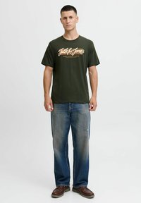 Olijfgroene katoenen t-shirt met witte en oranje tekst, gecombineerd met vervaagde blauwe baggy jeans en bruine veterschoenen.