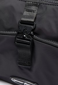 Podrobnosti blizu črne najlon torbe, ki vključuje plastično zaponko, tkano naramnico in šivani zadrgo na strani, kar prikazuje eleganten dizajn in funkcionalnost.