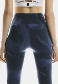 Kobieta w granatowych leginsach high-waisted w tie-dye i białym staniku sportowym, stojąca z ramionami swobodnie opuszczonymi wzdłuż ciała.