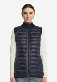 Marinblå pufferväst med horisontell quiltad design, hög krage och framdragkedja. Har en slät, glänsande yta och två fickor.