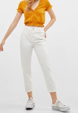 Femme portant un crop top à manches courtes et boutons de couleur jaune moutarde, un pantalon blanc taille haute retroussé et des baskets à plateforme blanches.