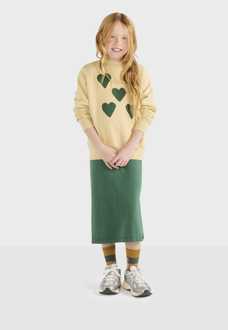 Cremefarvet sweatshirt med grønne hjerte designs, parret med en knælængde grøn nederdel. Accentueret med beige og grå sneakers. Langt, bølget hår.