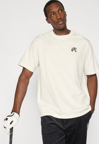 Κρέμ βαμβακερό μπλουζάκι με στρογγυλή λαιμόκοψη και κοντά μανίκια, με ένα μικρό μαύρο κεντημένο λογότυπο στο αριστερό στήθος.