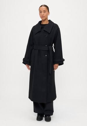 Melnā garā mētelī, kas izgatavots no teksturēta auduma, ir plaša apkakle, dubultie pogas un josta viduklī. Akcentēts ar platajām piedurknēm.