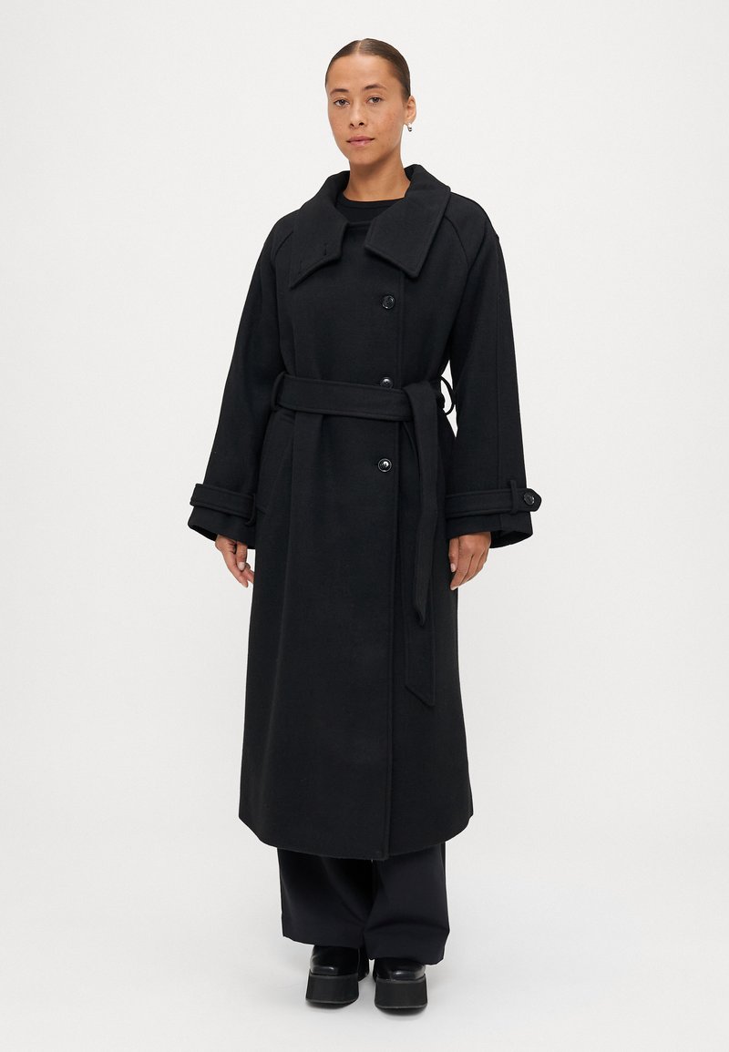 Melnā garā mētelī, kas izgatavots no teksturēta auduma, ir plaša apkakle, dubultie pogas un josta viduklī. Akcentēts ar platajām piedurknēm.