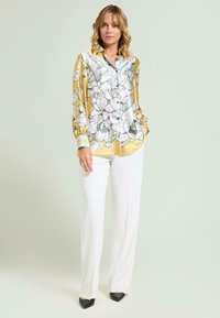 Silk blouse in yellow with floral print, featuring a collared neckline and button-down front, paired with white trousers and black pointed shoes.
