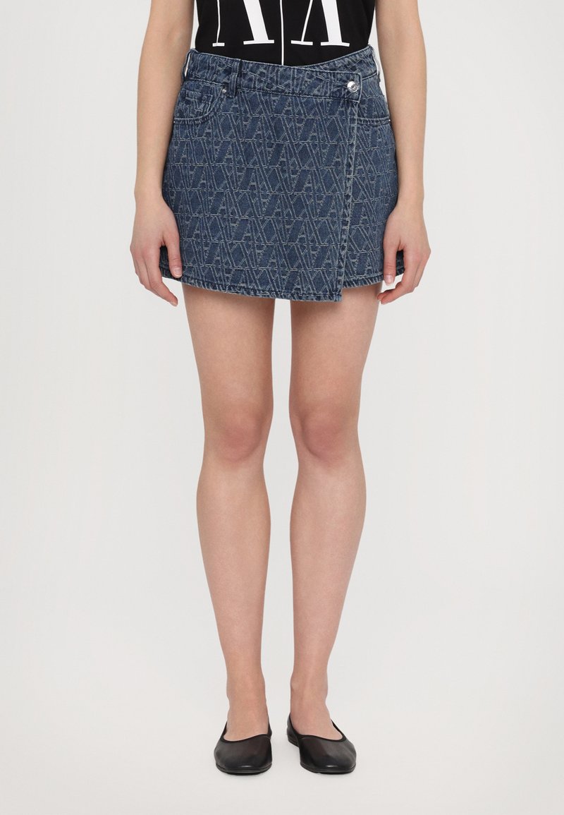 Jupe en jean courte avec un design croisé, de couleur bleu foncé et avec un motif subtil. Fini avec une fermeture à bouton sur le devant et des passants de ceinture.