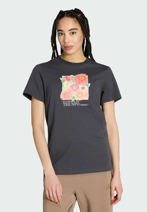 Young woman with braided hair wearing a black t-shirt with colorful floral square and "Flowers of Triumph" text, paired with beige pants.