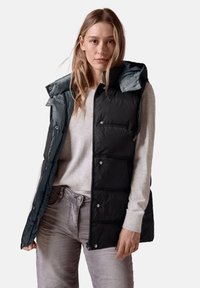 Schwarze Steppweste mit abnehmbarer türkisfarbener Kapuze, ausgestattet mit Druckknöpfen, Reißverschluss und seitlichen Taschen. Tragen über einem hellgrauen Oberteil.