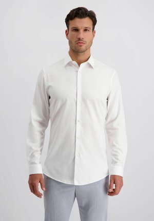 Man draagt een nauwsluitend wit overhemd met knopen en lichtgrijze broek, staand tegen een effen lichte achtergrond.