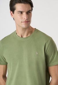 T-shirt verde oliva in morbido cotone, maniche corte, scollo a girocollo, con un piccolo logo ricamato sul petto. Texture liscia.