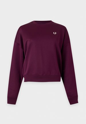 Un mannequin blond se tient devant un fond gris clair, vêtu d’un sweat-shirt bordeaux, d’un jean noir et de ballerines noires de style Mary Jane.