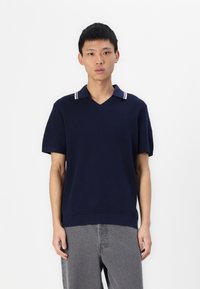 Polo din bumbac de culoare bleumarin, cu mânecă scurtă și guler în V, având accente albe cu dungi pe guler și un design tricotat texturat. Asociat cu pantaloni gri.