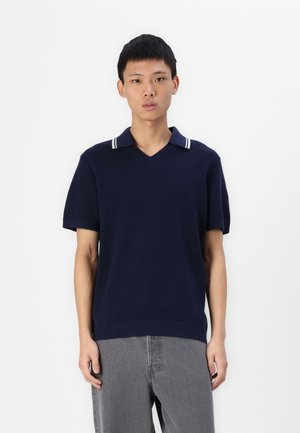 Mørkeblå polo-shirt med kortærmer og v-hals, prydet med hvide stribede detaljer på kraven og et struktureret strik design. Kombinationen er med grå bukser.