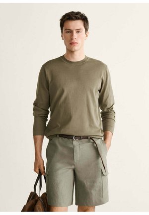 Jeune homme portant une chemise à manches longues vert olive et un short cargo assorti, tenant un sac marron dans une main, debout devant un fond uni.