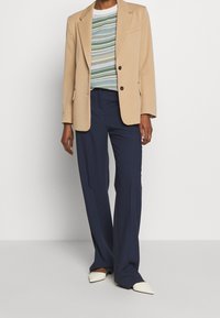 Beige blazer met twee knopen, gelaagd over een gestreept trui in blauw, groen en geel, gecombineerd met navy broek en witte puntige schoenen.