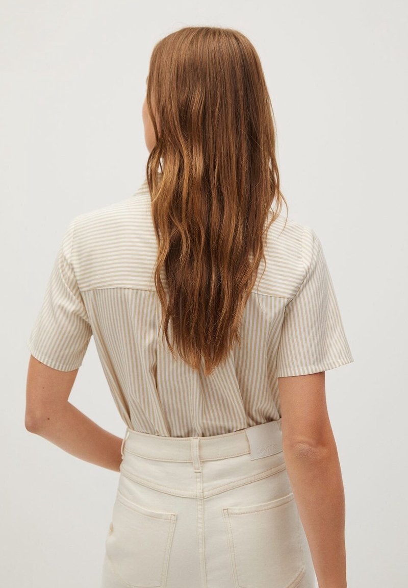 Personne aux longs cheveux bruns ondulés, portant une chemise beige et blanche rayée à manches courtes, rentrée dans un pantalon taille haute blanc cassé, de dos.