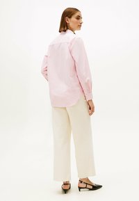 Blusa a rayas de color rosa claro, corte relajado, puños abotonados, y un dobladillo ligeramente más corto en la parte delantera. Combinada con pantalones crema y tacones negros tipo slingback.