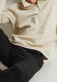 En person som bär en beige tröja med kvart dragkedja och svarta byxor, sitter med ena knät böjt inomhus mot en vit bakgrund.