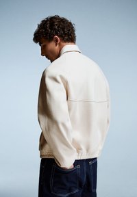 Jachetă din fleece de culoare crem, cu guler, cusături vizibile și manșete elastice, asociată cu pantaloni din denim întunecat. Vizualizare din spate.